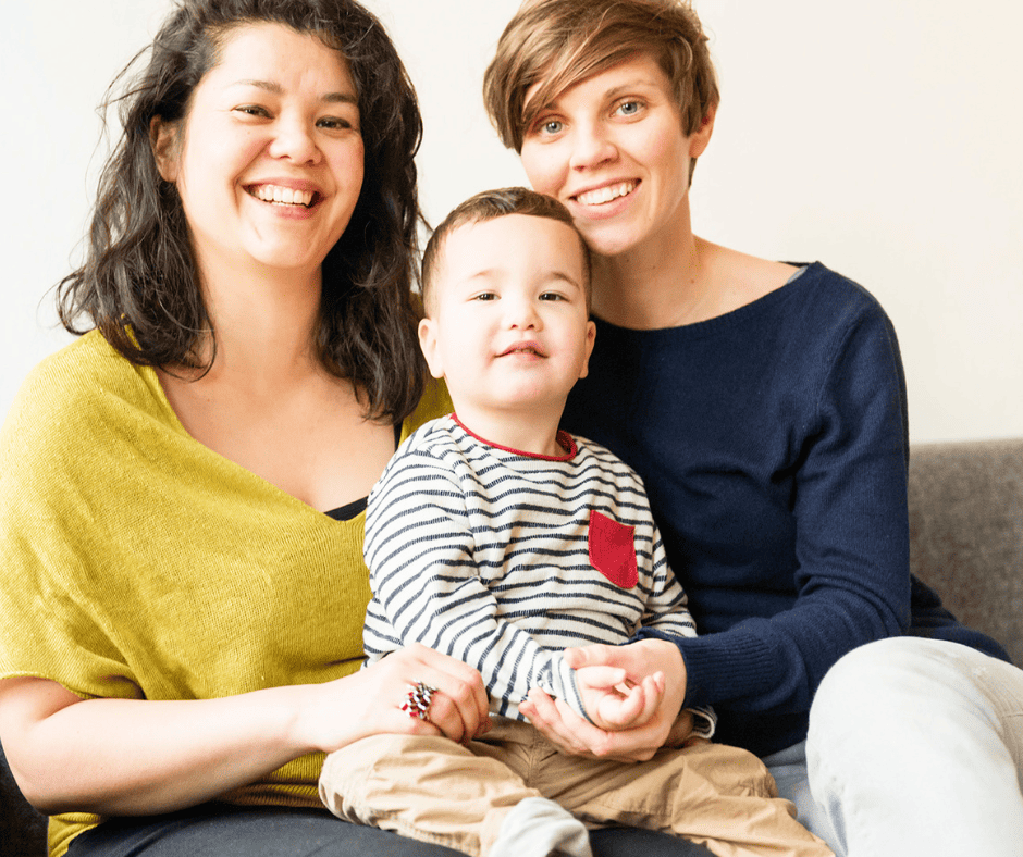 two mums and toddler sitting on sofa smile at camera